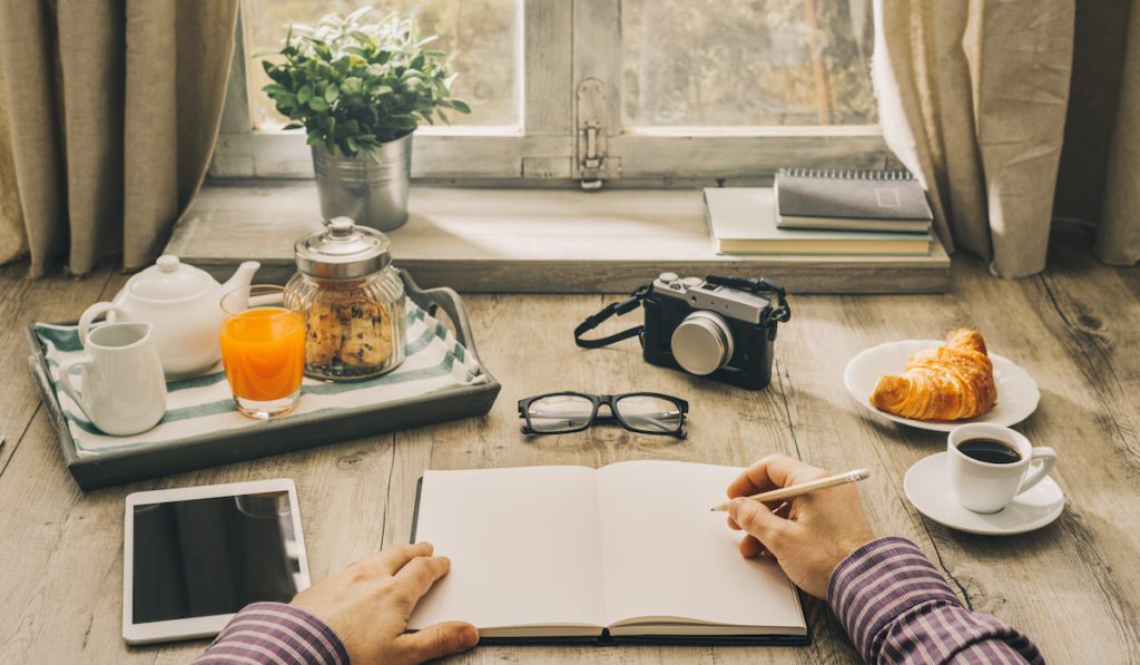 Man writing a journal in front of window with coffee, drinks, food, camera, glasses.
