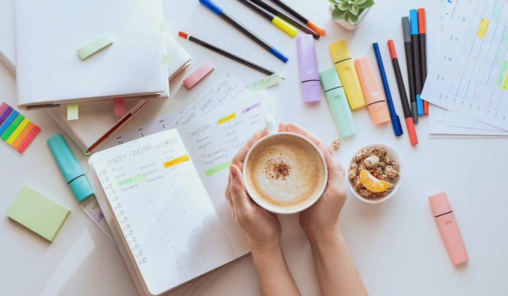 Top view of a white table with sticky notes, highlighter and hands with a cup of coffee. 