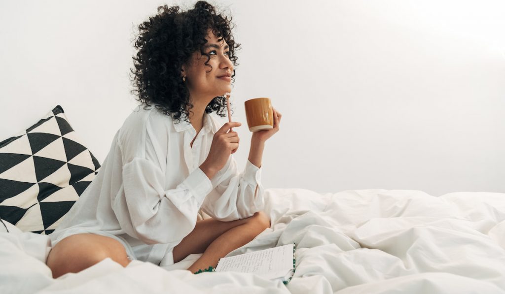  woman sitting on bed having coffee and writing on her journal. 